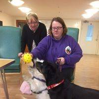 Kay is standing behind Lou, who is sat down playing with a black dog and a white dog.
